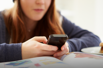 Una adolescentes mira el celular sentada en una mesa en la que hay libros.